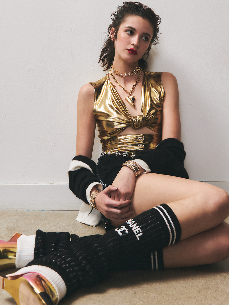 Model leaning aginst a white wall with bright red lip and gold top looking awar from the camera